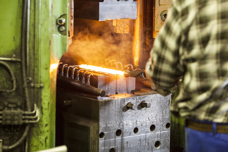 Ein Mann von hinten mit grün-kariertem Hemd hält mit einer Schmiedezange ein glühendes Bauteil, das auf dem Untergesenk der Schmiede liegt - Schmiedetechnik bei RÜBIG